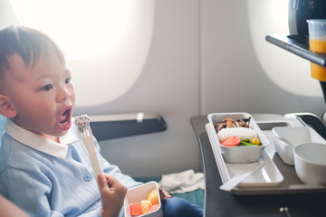 Cute little Asian 2 -3 years old toddler baby boy child wearing blue sweater eating food during flight on airplane. Flying with children, Happy air travel with kids & little traveler concept