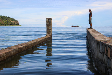 White Caucasian male traveler in sportswear standing on a stone pier on the longest lake in the world Tanganyika. In the background sailing boat with fishermen.