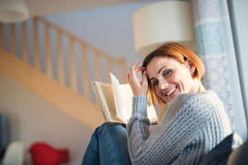 A young woman sitting indoors on a sofa at home, reading a book.