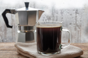 A cup of coffee on the background of geyser coffee machine and window with raindrops. Closeup, selective focus, wooden surface