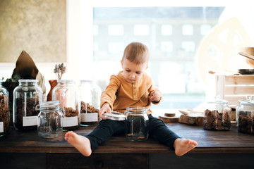 A barefoot small toddler boy sitting on a table in zero waste shop.