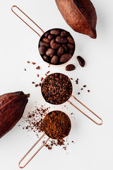Rose gold measuring cups of cocoa beans, cacao nips, cocoa powder and cocoa pods on a white...