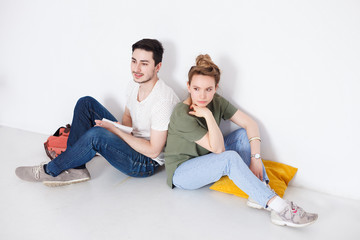 Indoor shot of happy young European couple freelancers relaxing at home after work day,sitting on floor at white wall, dressed jeans and casual t-shirt, smiling at camera. Lifestyle and people concept