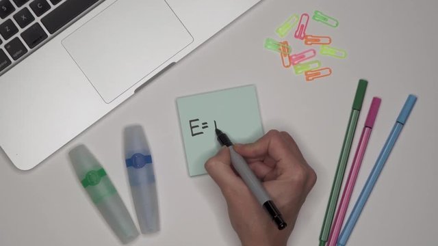 Womans hand writing E=mc2, einsteinian theory of relativity on adhesive notepad.