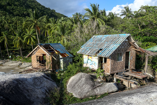 Old decaying bungalow type houses on beach rocks with palm trees in an abandoned tourist resort in Koh Phangan Thailand