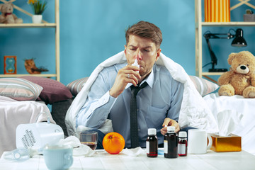 Bearded sick man with flue sitting on sofa at home covered with warm blanket and using nasal spray....
