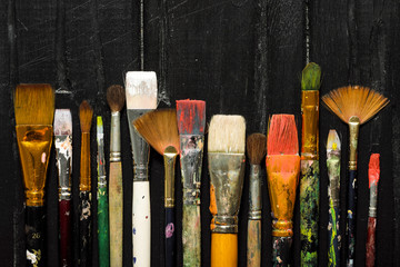 Artist's tools, brushes in paint are in a row on a black wooden background.