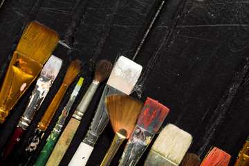 Artist's tools, brushes in paint are in a row on a black wooden background.
