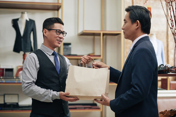 Fototapeta na wymiar Smiling tailor giving customer paper-bag with bespoke suit