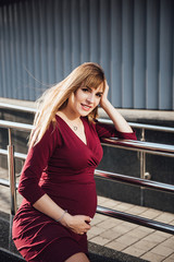 Pregnant girl blonde in burgundy dress. In the big city. Harm to the environment. Long hair. 9 months in anticipation. Happiness to be a mom. Happiness. Portrait.