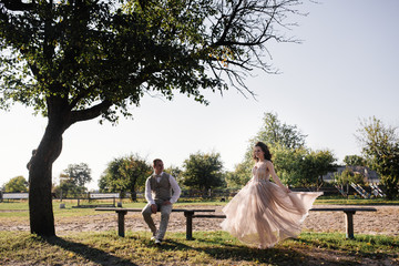 Wedding at sunset. Couple is sitting on a bench under a tree. Beige dress with sparkles. Light suit with a bow tie. The bride and groom embrace and kiss.