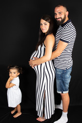 Handsome bearded man and his beautiful pregnant wife are hugging and smiling with little child girl daughter while standing on black background