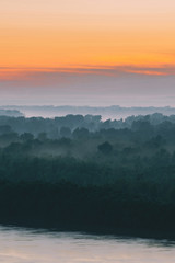 Fototapeta premium Mystical view on riverbank of large island with forest under haze at early morning. Mist among layers from tree silhouettes under warm predawn sky. Morning atmospheric landscape of majestic nature.