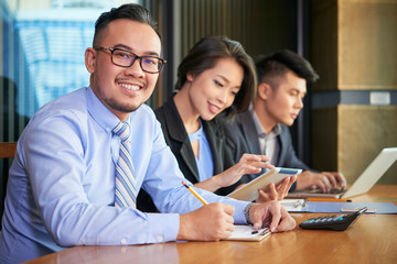 Cheerful Asian entrepreneur filling document when working at tablet with his colleagues