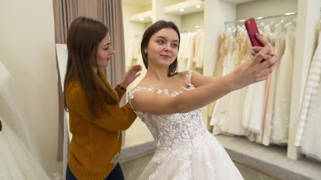 A future bride takes selfie with a bridesmaid in a fitting room while trying on a new wedding dress. A bridesmaid fixes bride's hairstyle and creates a ponytail by pulling all hair to the back.