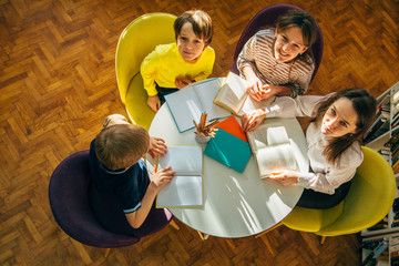 Teacher and children looking up at camera while sitting in a circle round table at library. Top view of librarian sitting with four children. Teacher reading book to girl and boys at school