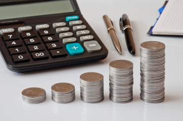 Coin columns, calculator, notepad and pen on a white table