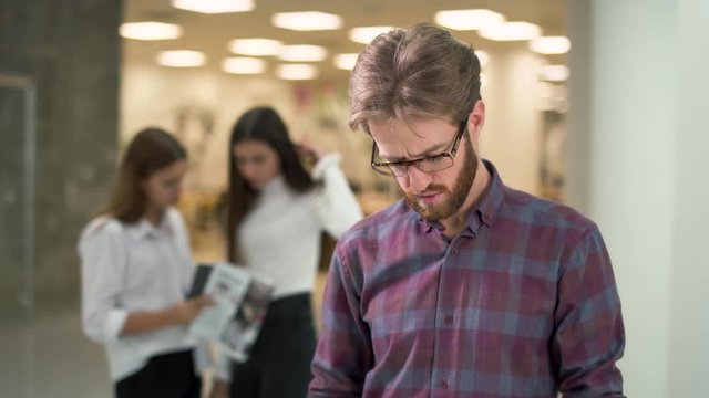 Portrait a guy with a beard in glasses and casual plaid shirt reading documents with sales reports standing in the lobby of an office business center. Two girls colleagues on background.