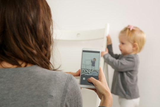 Mother taking a photo of her baby girl with a cell phone in a nursery room