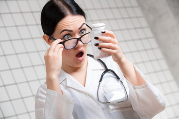 Surprised female doctor gesturing with hands and pills while standing with mouth open on medical office background.