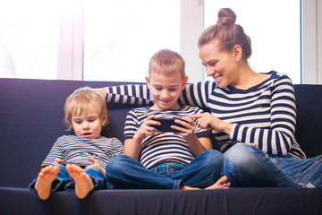 Obraz premium mother with two brothers blond boys on a blue sofa at home playing smartphones