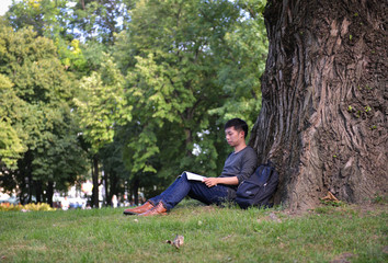 young male guy of asian appearance student sitting under a tree with a book and laptop, learn college and university backpack