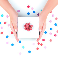 Obraz na płótnie Canvas Hands giving or receiving a gift box with red bow flower over multicolored confetti. Present and celebration isolated