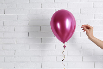 Young woman piercing bright balloon near brick wall, space for text