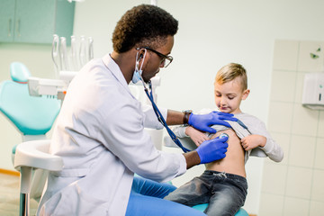 Obraz premium Physician looking at patient and examining posture of little boy. Happy kid smiling, looking away. African American Doctor having stethoscope on neck, wearing in white medical gown