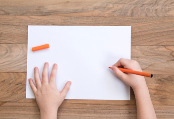 Child's hands with orange felt-tip on the wooden table with a piece of paper. Top view. 