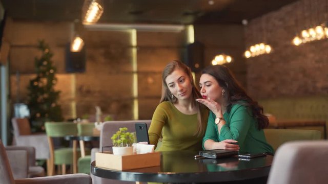 Two female friends are sitting in a cafe indoors and making a lot of selfies with a different facial expressions. Both women are wearing green blouses.