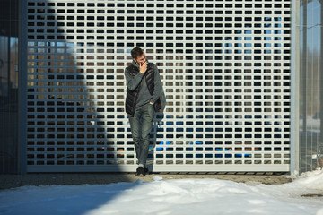 A young handsome guy in a black vest is smoking a cigarette in front of a metal gate on the street in spring.