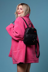 Blonde cheerful girl posing in stylish pink coat in studio with black backpack