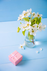 Little gift box and bouquet of tender white flowering branches in glass vase on blue wooden background.