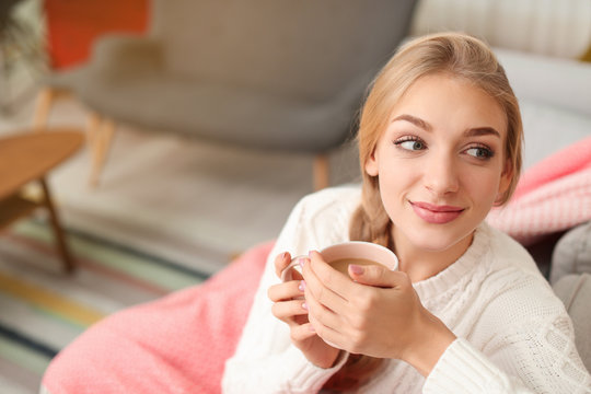 Beautiful young woman wrapped in plaid sitting on sofa with cup at home. Winter atmosphere