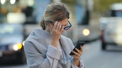 Beautiful blonde woman using a smartphone and fixing her hair while standing on the street in downtown. Close-up portrait. Blurred background. People passing by.