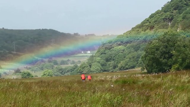 Two hikers trekking through a green landscape with a rainbow above them
