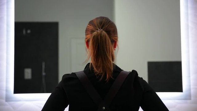 Cute blonde woman in black shirt is wearing leather belt. Looking in the mirror while standing on light grey background.