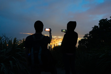 Fototapeta na wymiar Two tourists using cameras on mobile devices/phones to photograph urban night lights from top of landmark