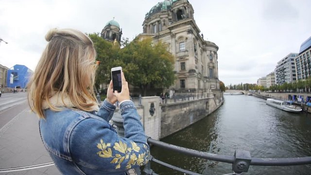Profile of pretty blonde woman taking panorama pictures of wonderful cityscape on a smartphone over the riverbank.