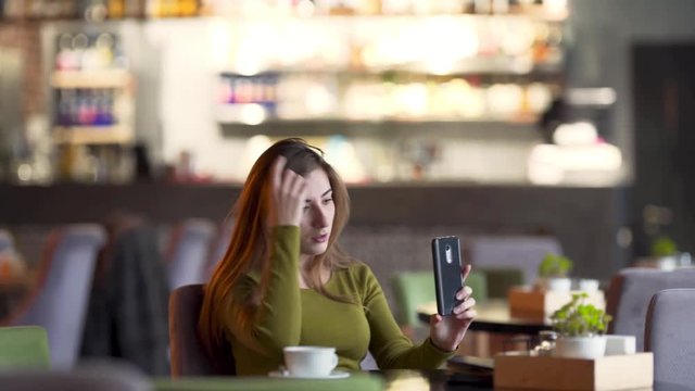 Portrait of a young charming woman who is  sitting in a cafe indoors and talking in a video chat with her friend. She is looking absolutely relaxed and easy.