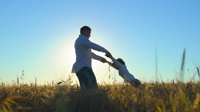 silhouette of father and son playing and enjoying sunset in wheat field in nature on summer day. happy family walking outdoors, having fun. father circling and turns little child boy by taking hands