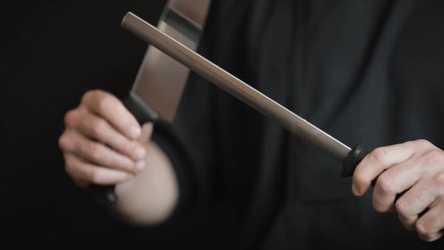 Close-up of male hands using a honing steel rod to sharpen a knife blade on a black background