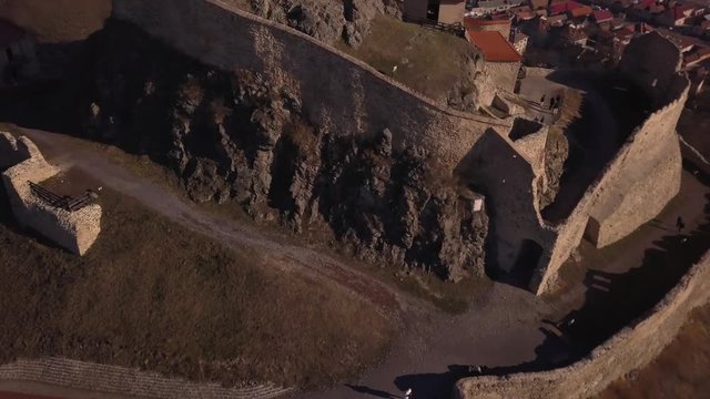 Drone rotates and descends giving an exterior view of the medieval architecture and ruins of Rupea Fortress. High stone walls and buildings perched on a hilltop.