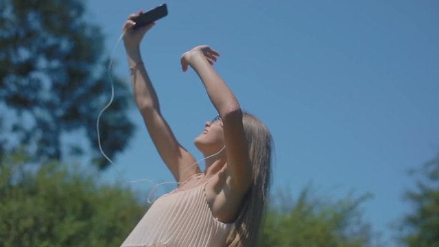 slow motion blond girl with long loose flowing hair in earphones holds smartphone dancing in green park under blue sky