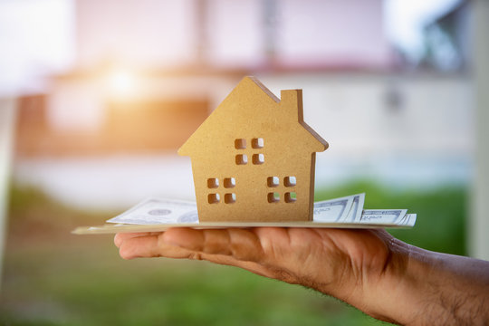 Hand A Businessman Investing And Saving Money Or Borrowing Money From Financial Institutions To Buy Home For The Future Is A Valuable Asset In The Concept Of House Model Coupled With Dollar Banknotes.