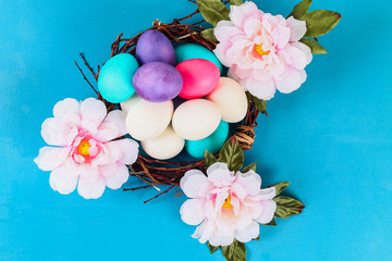 Easter colored multicolored eggs lying inside the nest next to the flowers on a blue background.