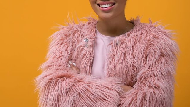 Modern female hipster in pink fur coat standing yellow background crossing hands