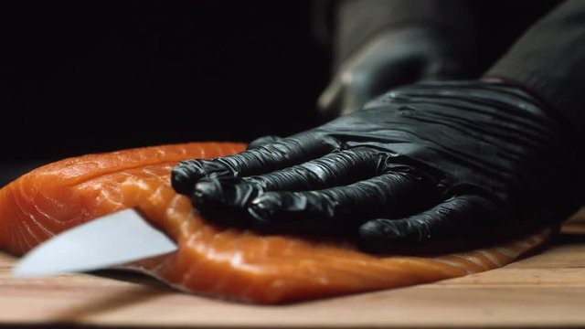 Close-up chef with piece of red salmon. Professional chef in black gloves cuts piece of fresh salmon into thin slices with sharp knife