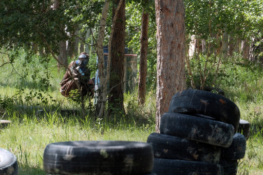 Paintball - Team game. A teenager in camouflage clothing with a weapon in his hands awaits opponents in an ambush in the forest among trees, iron barrels, automobile tires.